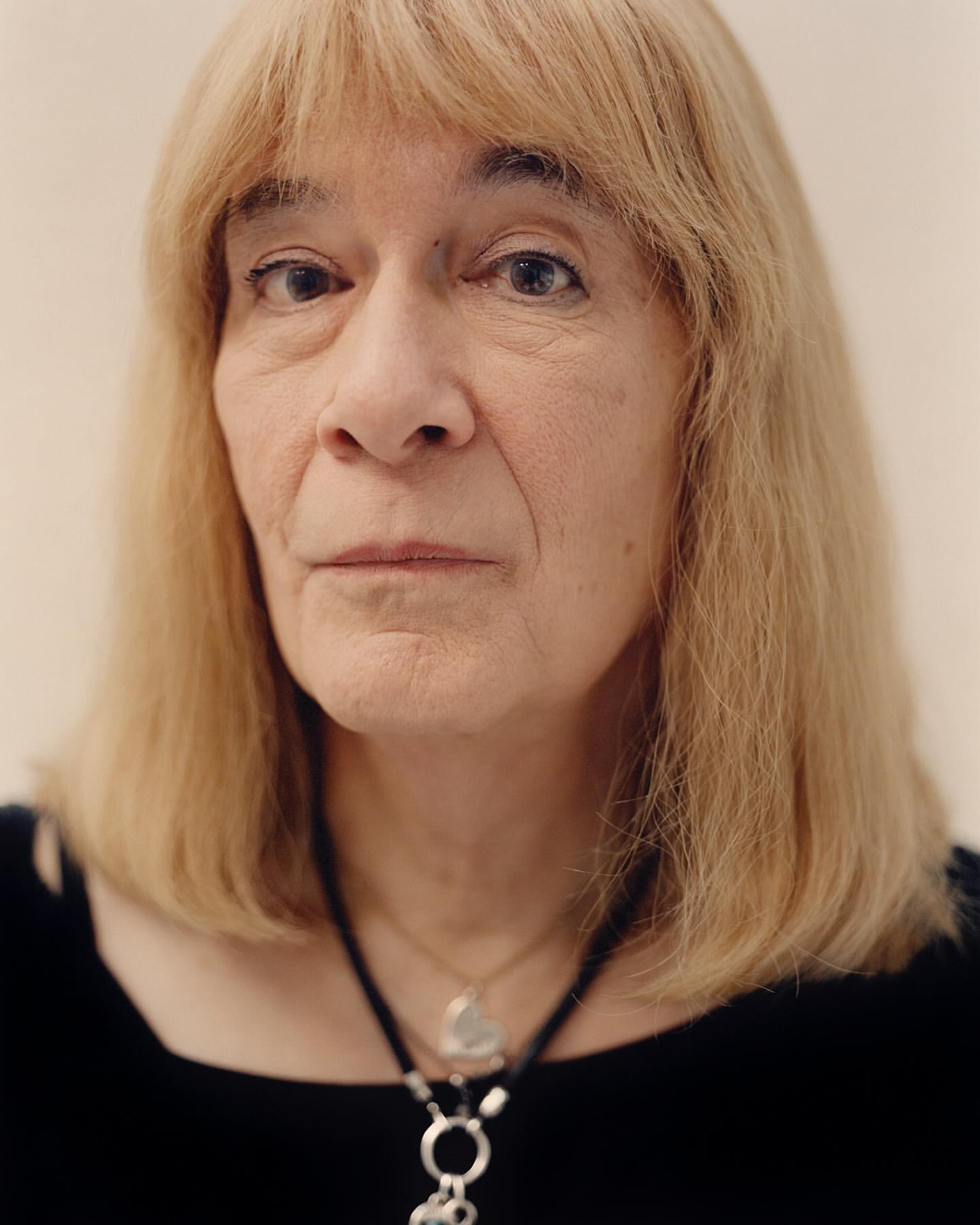 Close-up portrait of a mature adult female with blond hair wearing a black top and necklace, set against a light background.