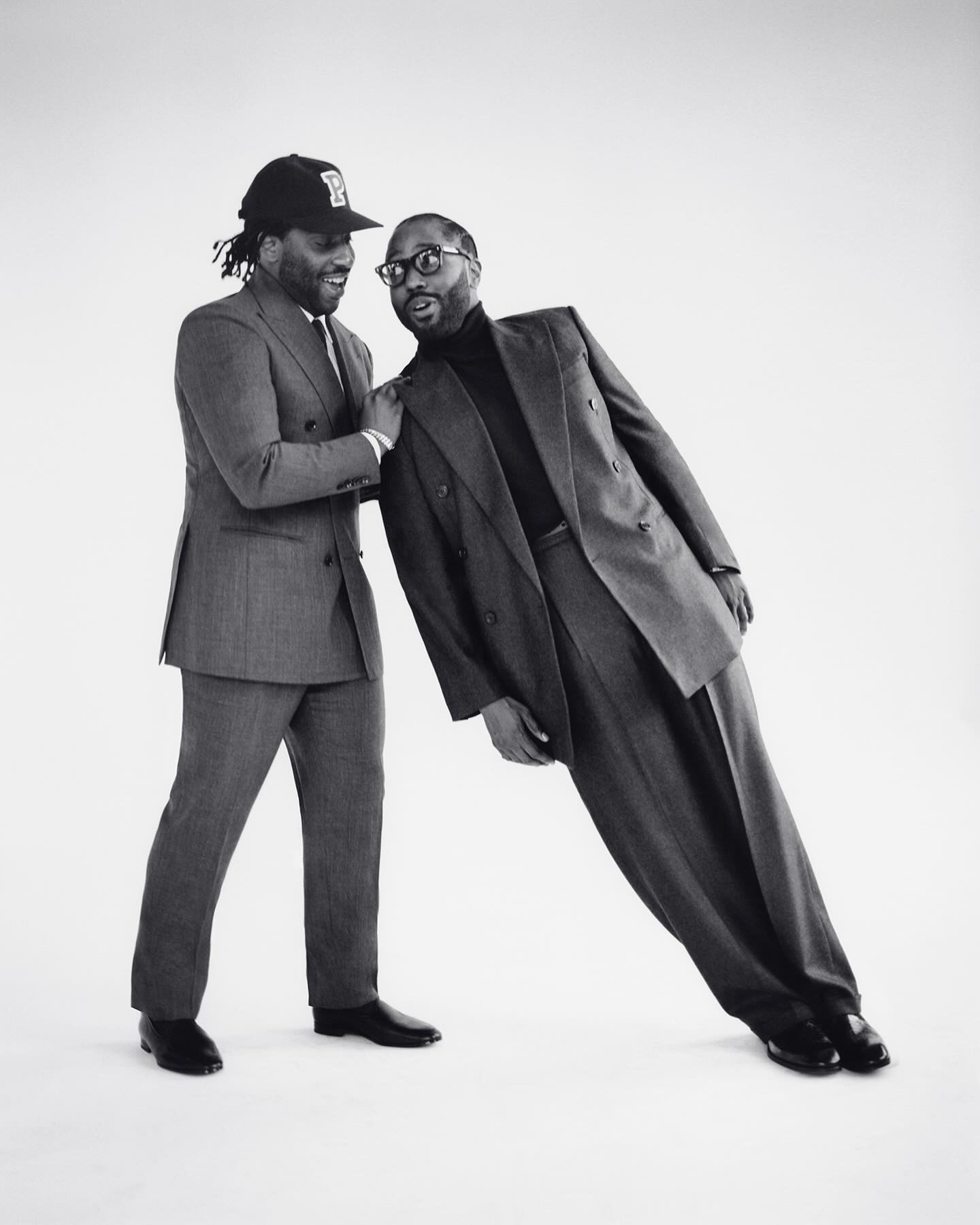 Two adult men in suits pose in a bright studio. One man leans on the other, who wears a baseball cap and smiles, against a white backdrop.