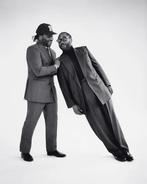 Two adult men in suits pose in a bright studio. One man leans on the other, who wears a baseball cap and smiles, against a white backdrop.