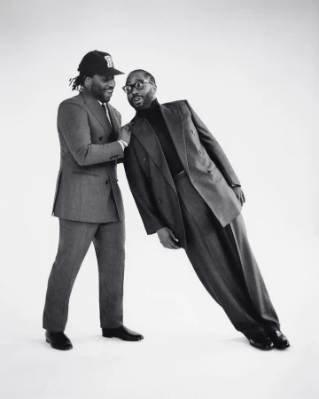 Two adult men in suits pose in a bright studio. One man leans on the other, who wears a baseball cap and smiles, against a white backdrop.