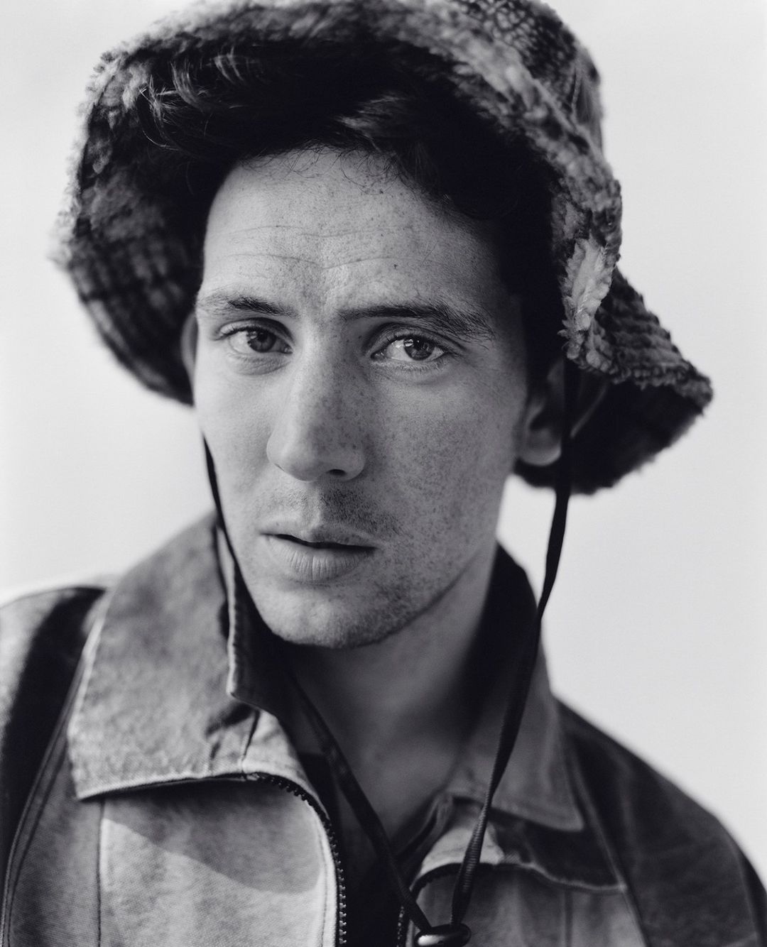 Close-up portrait of a man wearing a bucket hat and jacket against a plain background