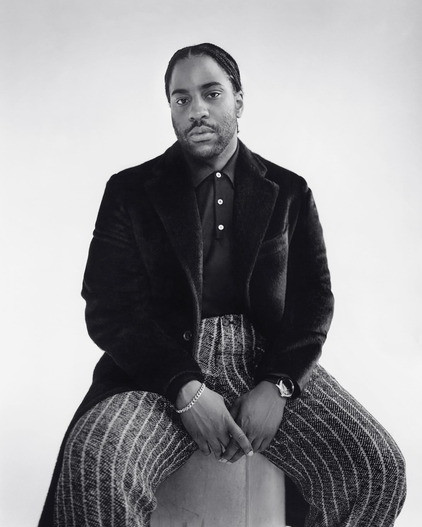 Monochrome portrait of an adult male sitting on a stool, wearing a black coat, black polo shirt, and vertically striped trousers, against a white backdrop.