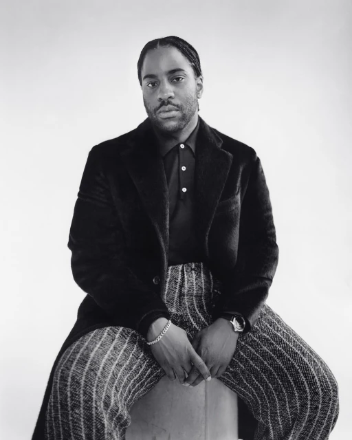 Monochrome portrait of an adult male sitting on a stool, wearing a black coat, black polo shirt, and vertically striped trousers, against a white backdrop.