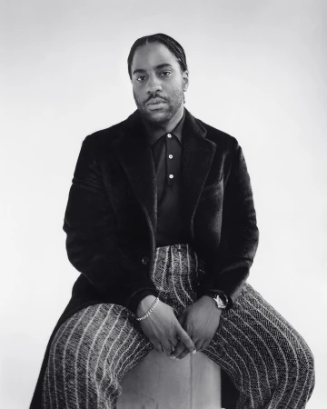 Monochrome portrait of an adult male sitting on a stool, wearing a black coat, black polo shirt, and vertically striped trousers, against a white backdrop.