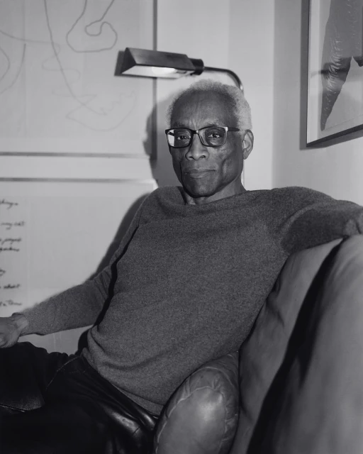 Black and white portrait of an African-American man wearing glasses and a grey sweater, sitting on a leather sofa with artwork in background.