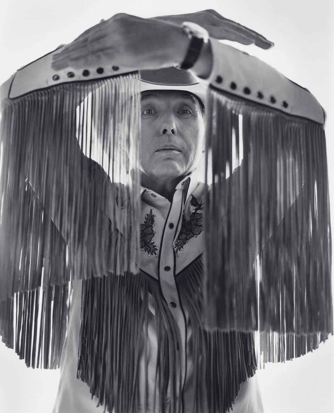 Monochrome portrait of a mature adult wearing a fringed western shirt and cowboy hat with hands raised overhead in a photo studio.