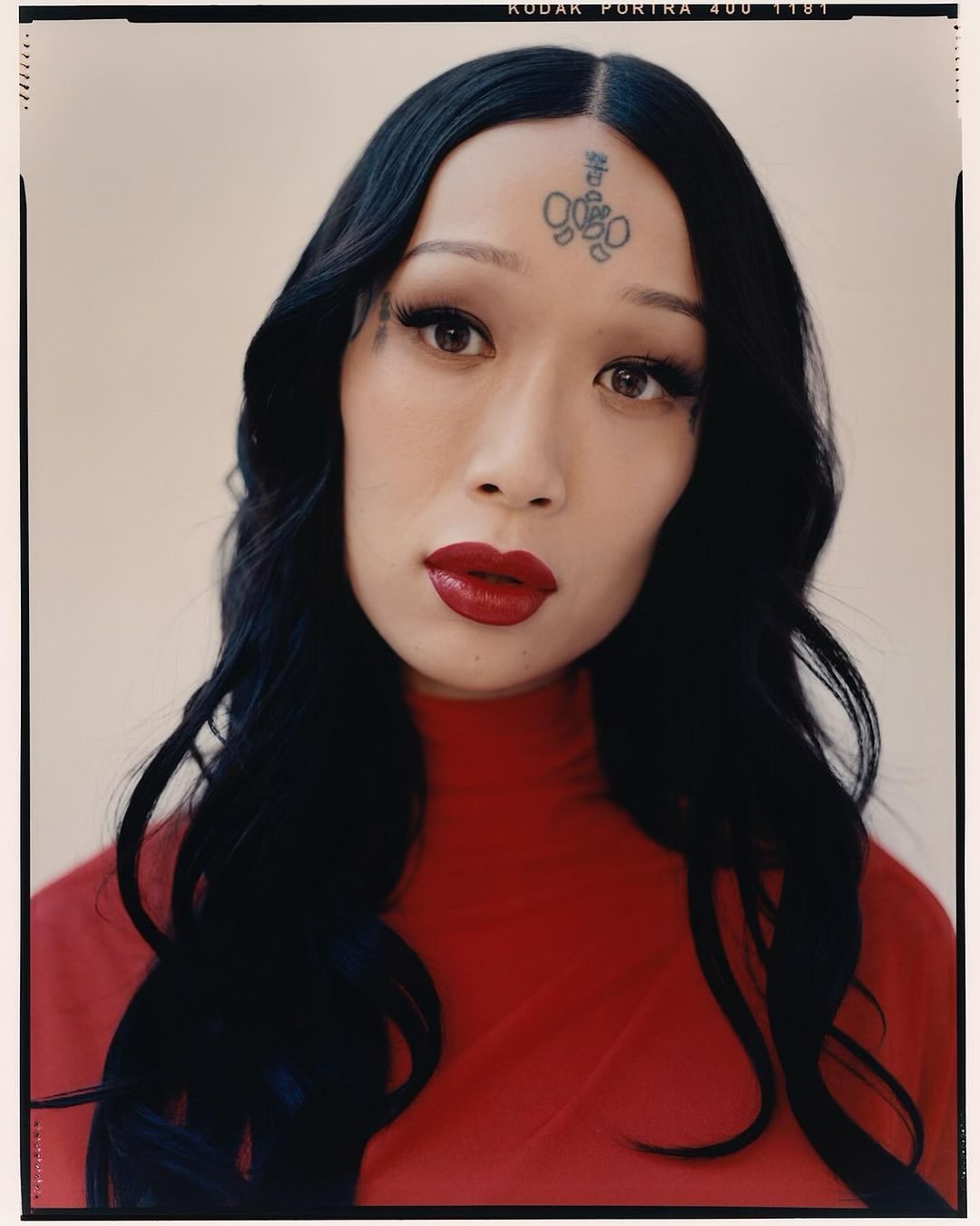 Glamour headshot of an adult woman with an intricate tattoo on her forehead, wearing red lipstick and a red turtleneck sweater.