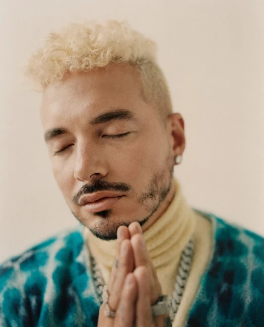 Portrait of an adult male celebrity, J Balvin, with his eyes closed, wearing jewelry and a patterned shirt, against a neutral backdrop.