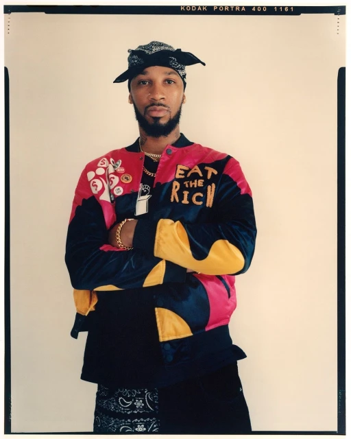Portrait of an adult male with crossed arms wearing a colorful jacket and bandana against a neutral background in an eye-level shot.