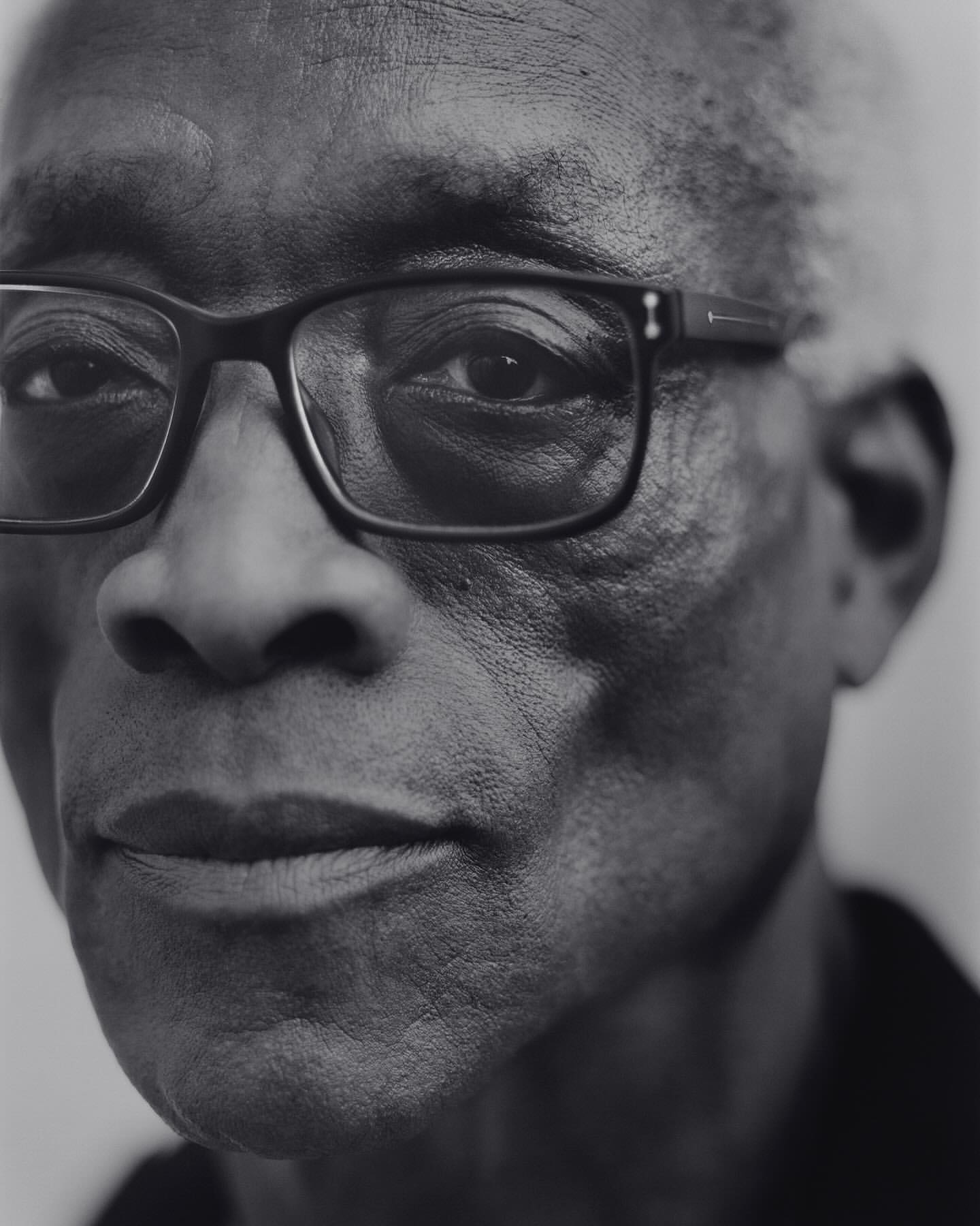 Monochrome portrait of an adult Black man with glasses, gazing directly at the camera with neutral expression, against a plain background.