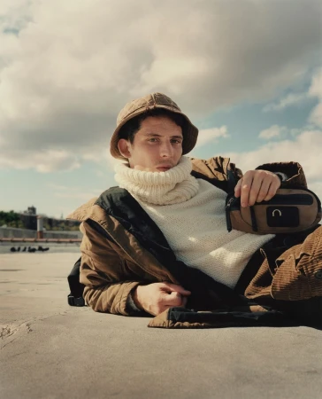 Man wearing a brown bucket hat and white turtleneck sweater lying outdoors under an overcast sky.