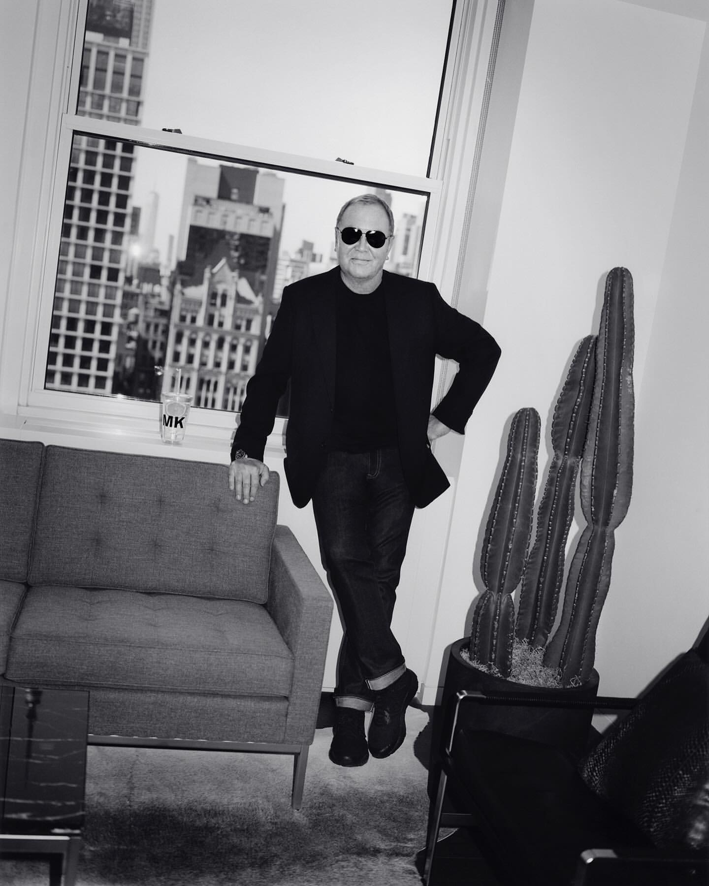 Black and white shot of an adult male wearing sunglasses, suit jacket, and jeans, posing indoors near a window overlooking a cityscape.