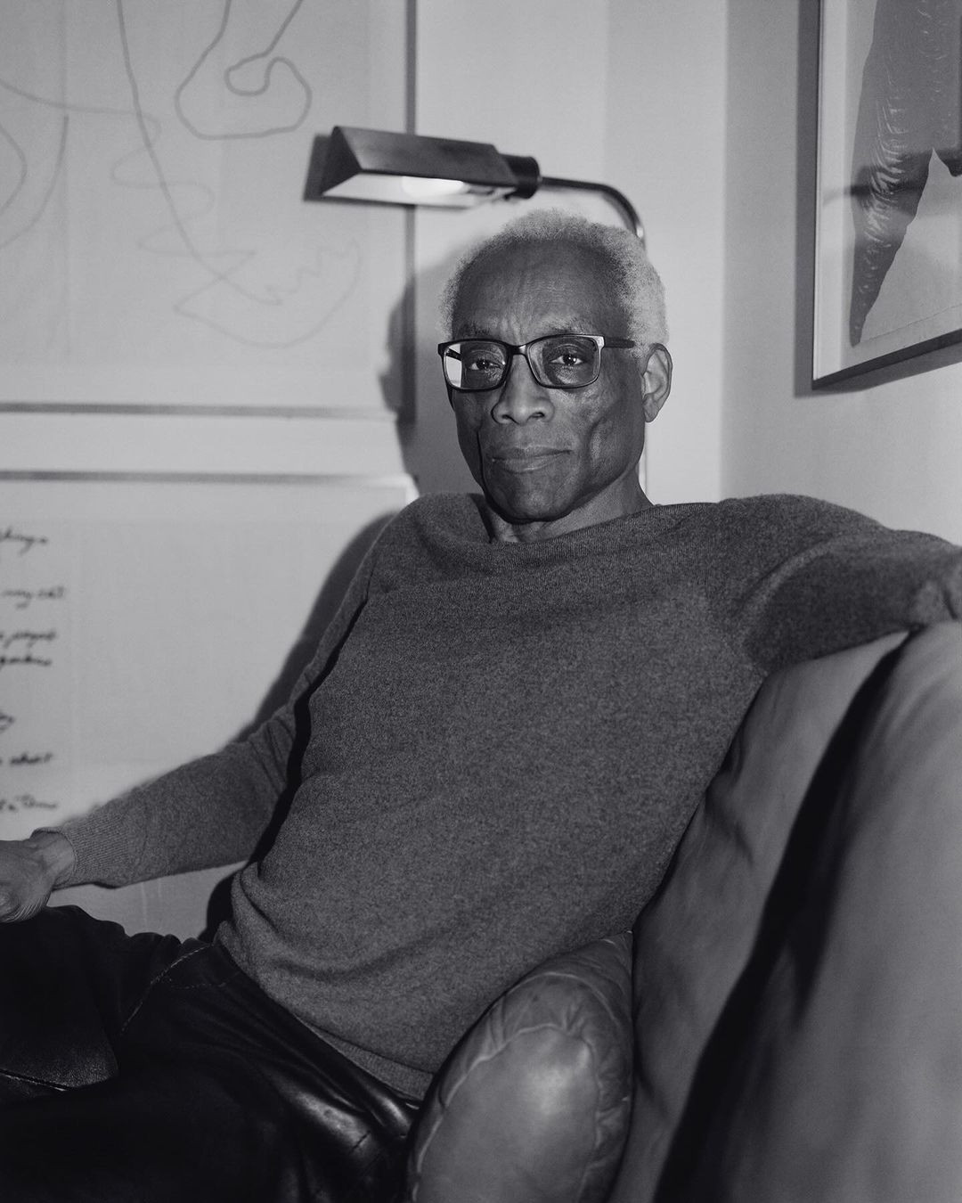 Monochrome portrait of an adult male with eyeglasses, gray sweater, and black leather pants, posed on a dark sofa in a room with artwork.