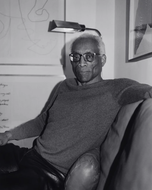 Monochrome portrait of an adult male with eyeglasses, gray sweater, and black leather pants, posed on a dark sofa in a room with artwork.