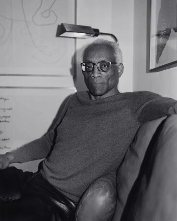 Monochrome portrait of an adult male with eyeglasses, gray sweater, and black leather pants, posed on a dark sofa in a room with artwork.