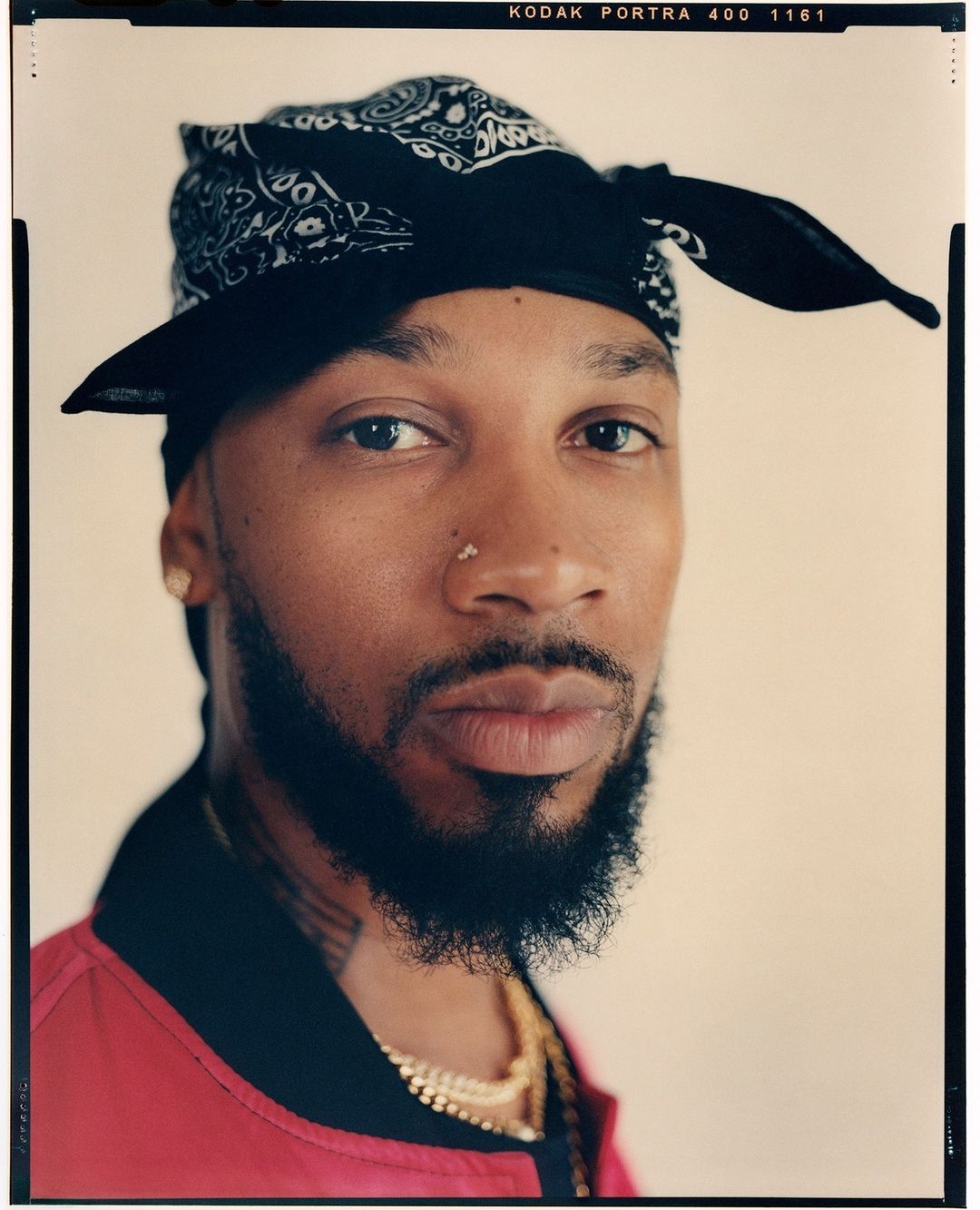 Close-up portrait of an adult male with a beard, wearing a black bandana and a red jacket, gazing intently at the camera.
