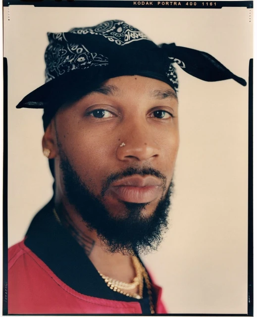 Close-up portrait of an adult male with a beard, wearing a black bandana and a red jacket, gazing intently at the camera.
