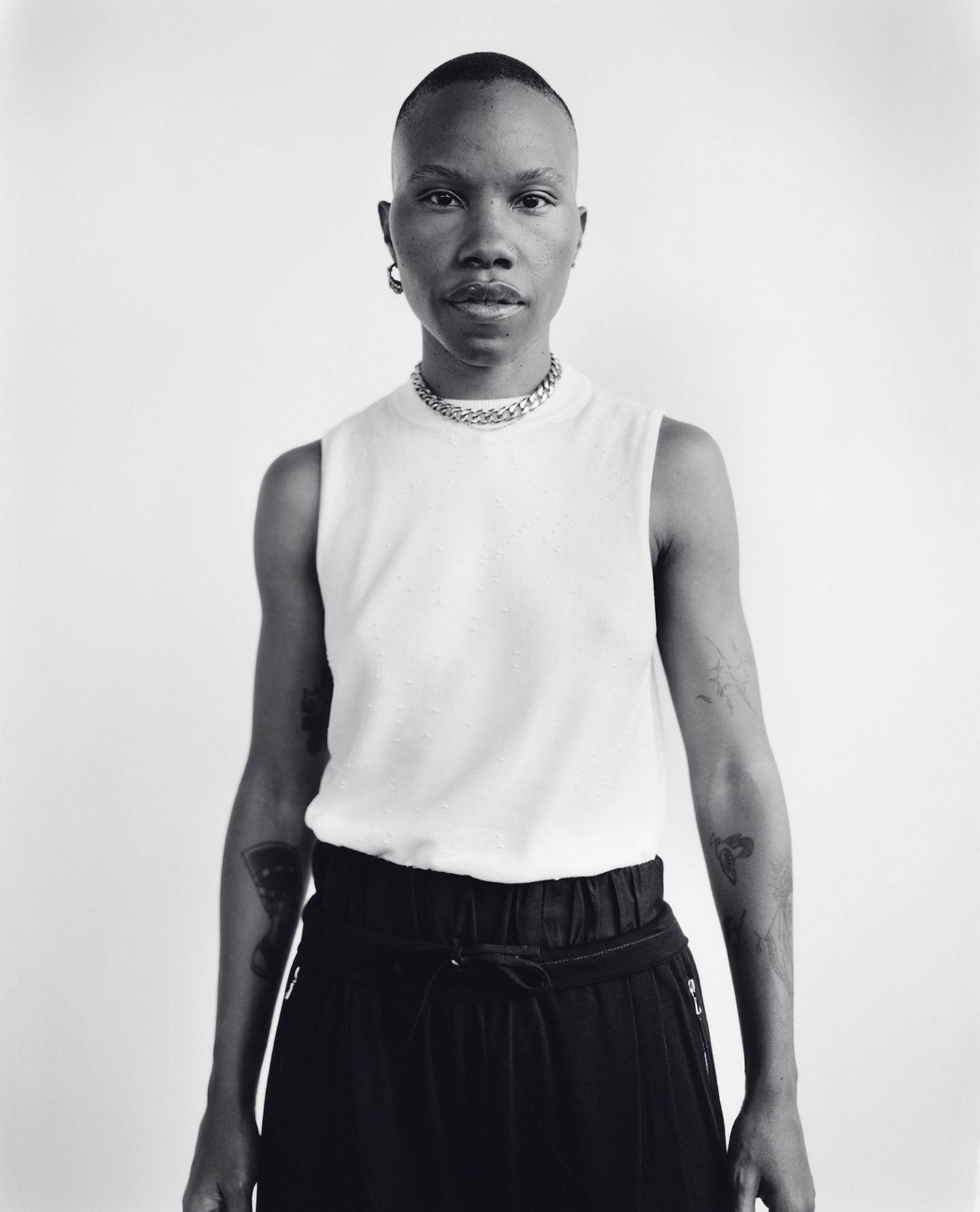 Portrait of a black person with a shaved head wearing a white tank top and dark pants against a plain background