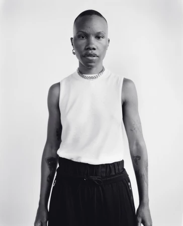 Portrait of a black person with a shaved head wearing a white tank top and dark pants against a plain background
