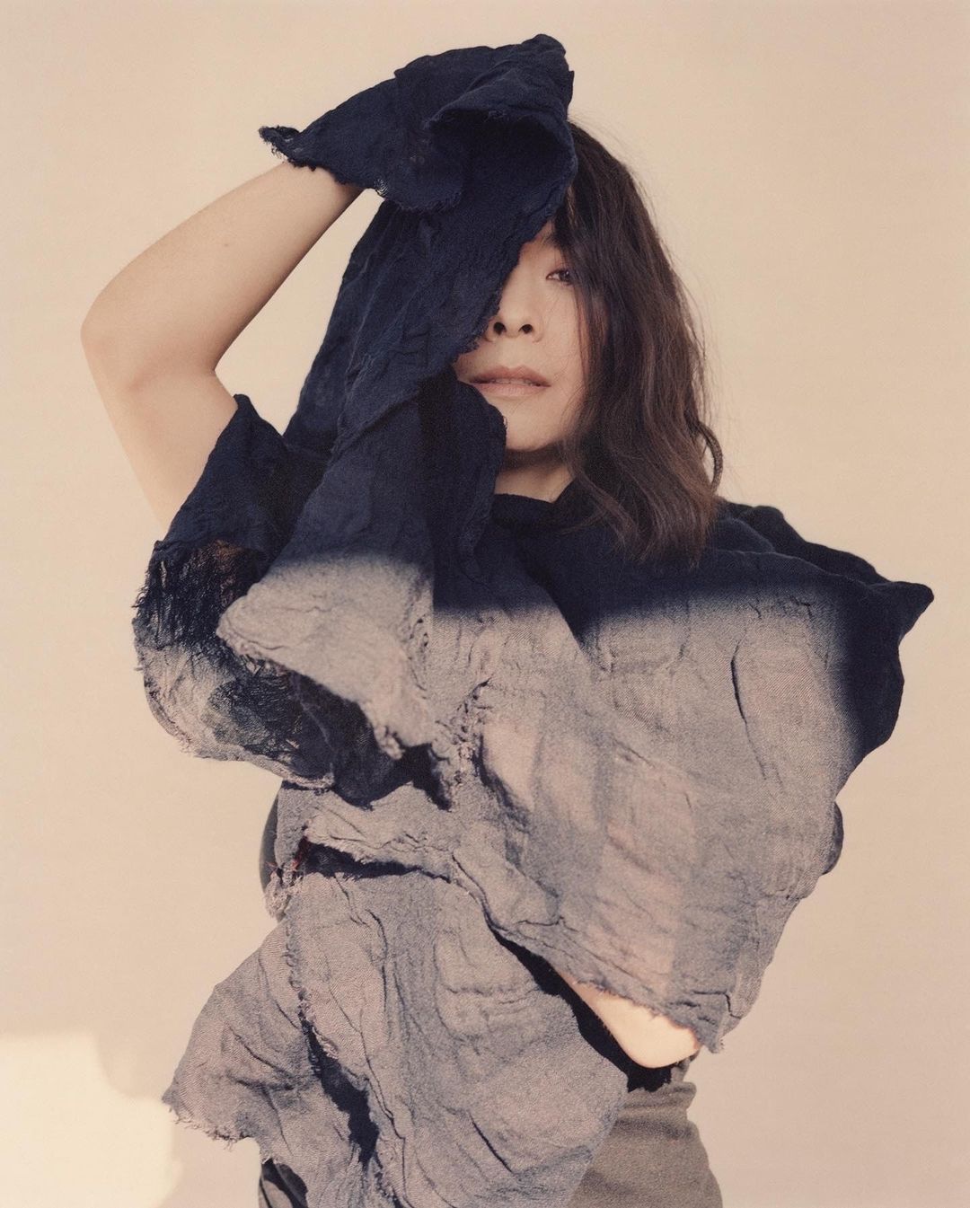 An adult female poses indoors with a flowing, layered fabric garment in blue and grey, partially covering her face in a studio portrait.