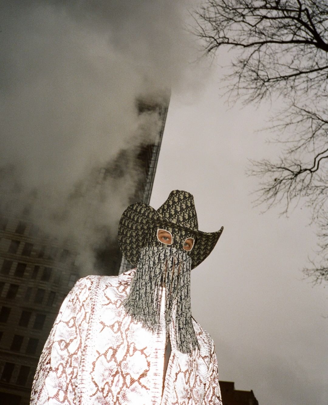 Fashionable individual wearing a patterned jacket and a matching Dior cowboy hat and mask standing outdoors with an urban backdrop.