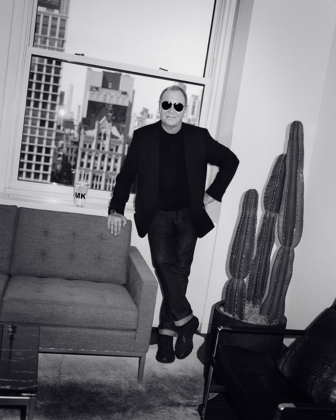 Black and white studio shot of a man in sunglasses posing by a window in a suit, near a couch and cactus plant.