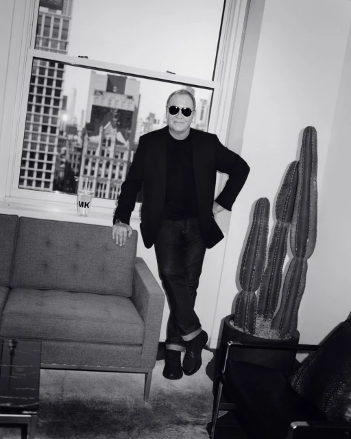 Black and white studio shot of a man in sunglasses posing by a window in a suit, near a couch and cactus plant.