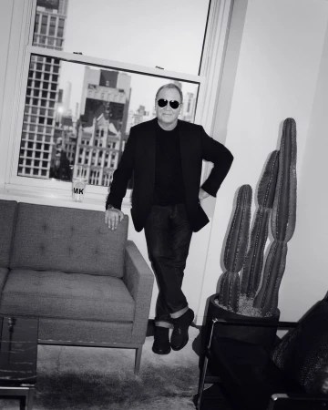 Black and white studio shot of a man in sunglasses posing by a window in a suit, near a couch and cactus plant.