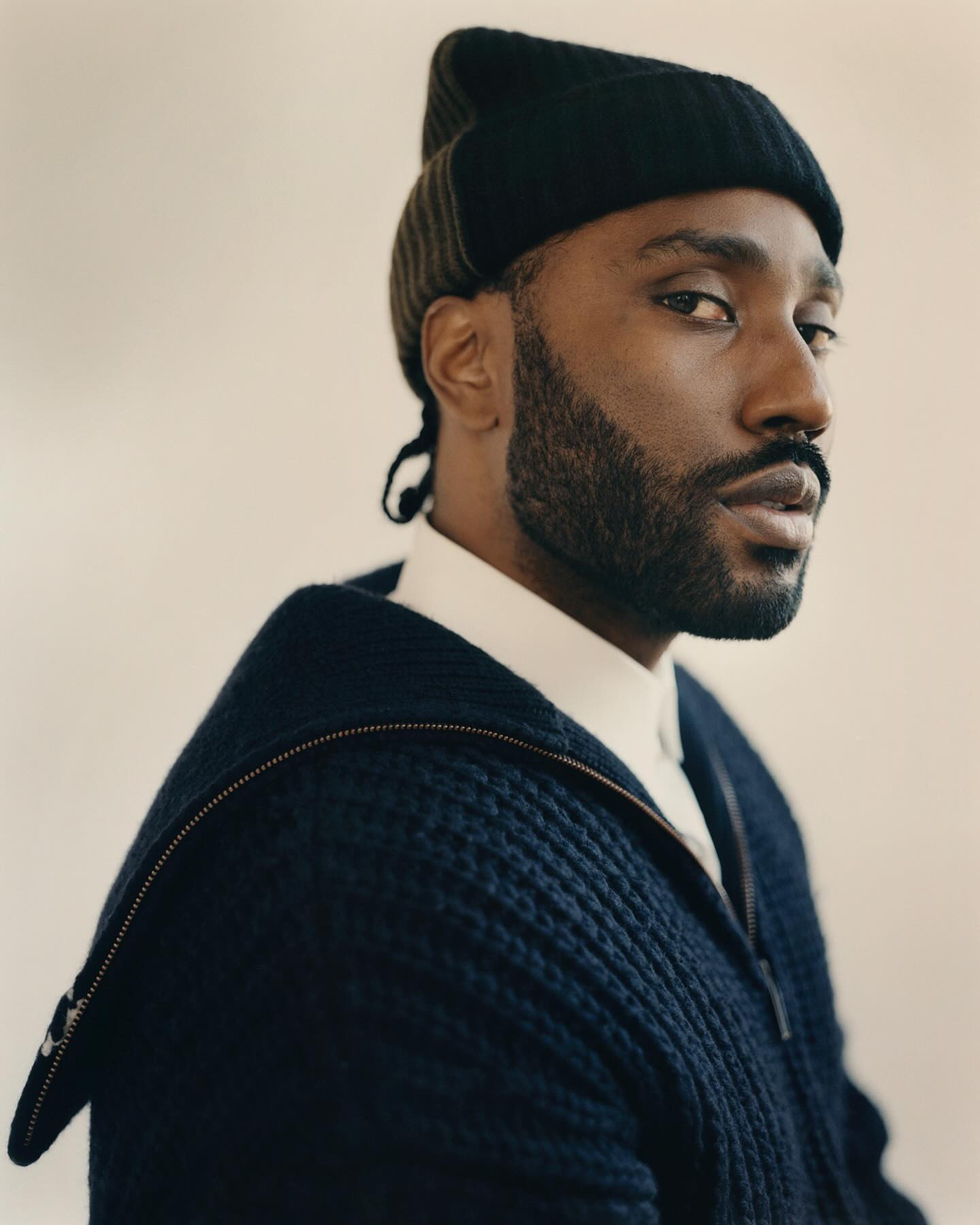 Portrait of a stylish Black man with a beard, wearing a navy blue zip-up cardigan and a black beanie, set against a plain background.