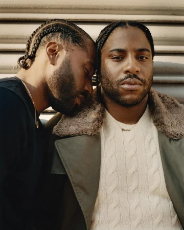 Close-up portrait of two Black men with braided hair leaning together, wearing layered winter clothing against a textured urban storefront background.