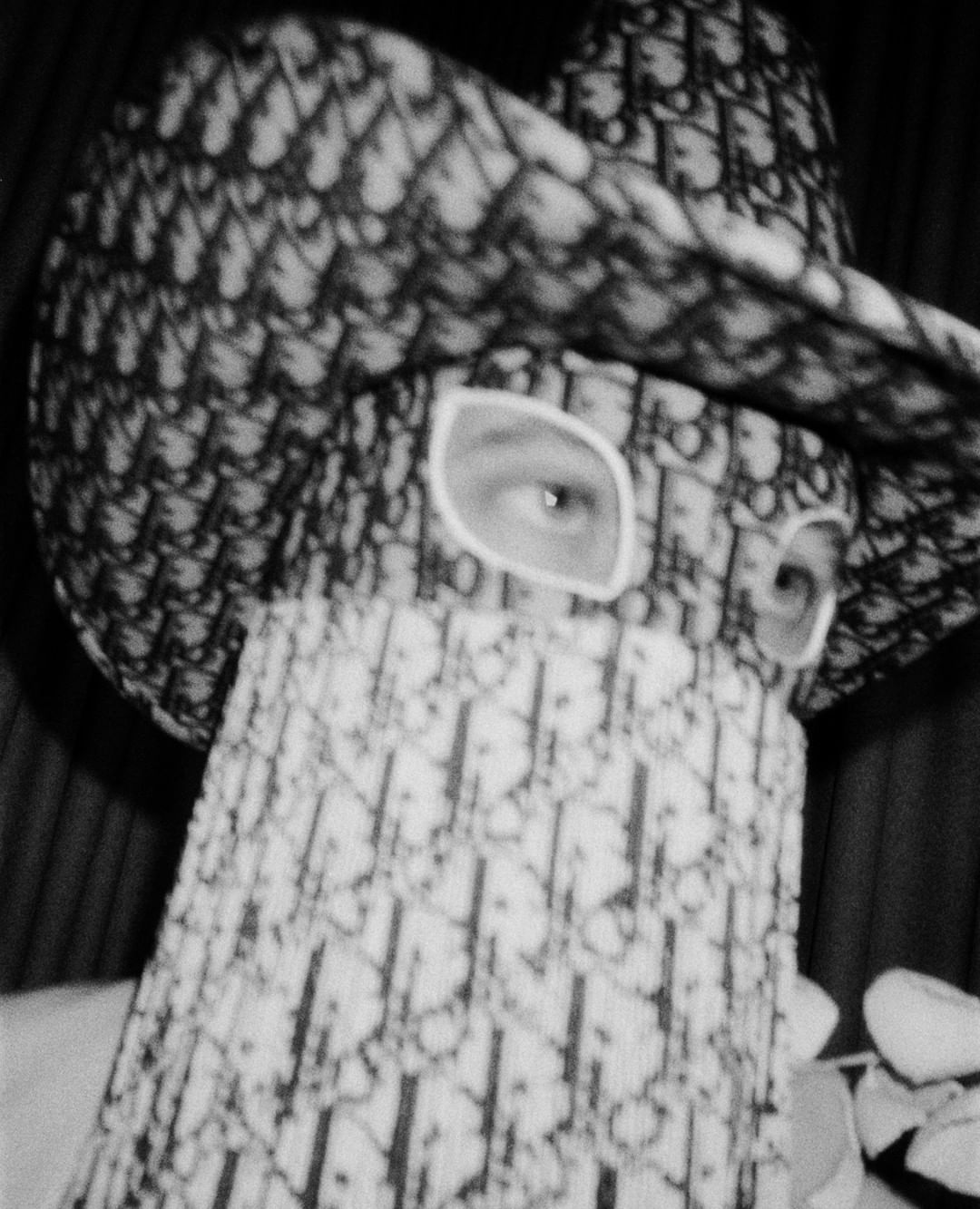 Person wearing a Dior-patterned hat and face covering, only eyes visible, in a high-contrast black and white close-up.