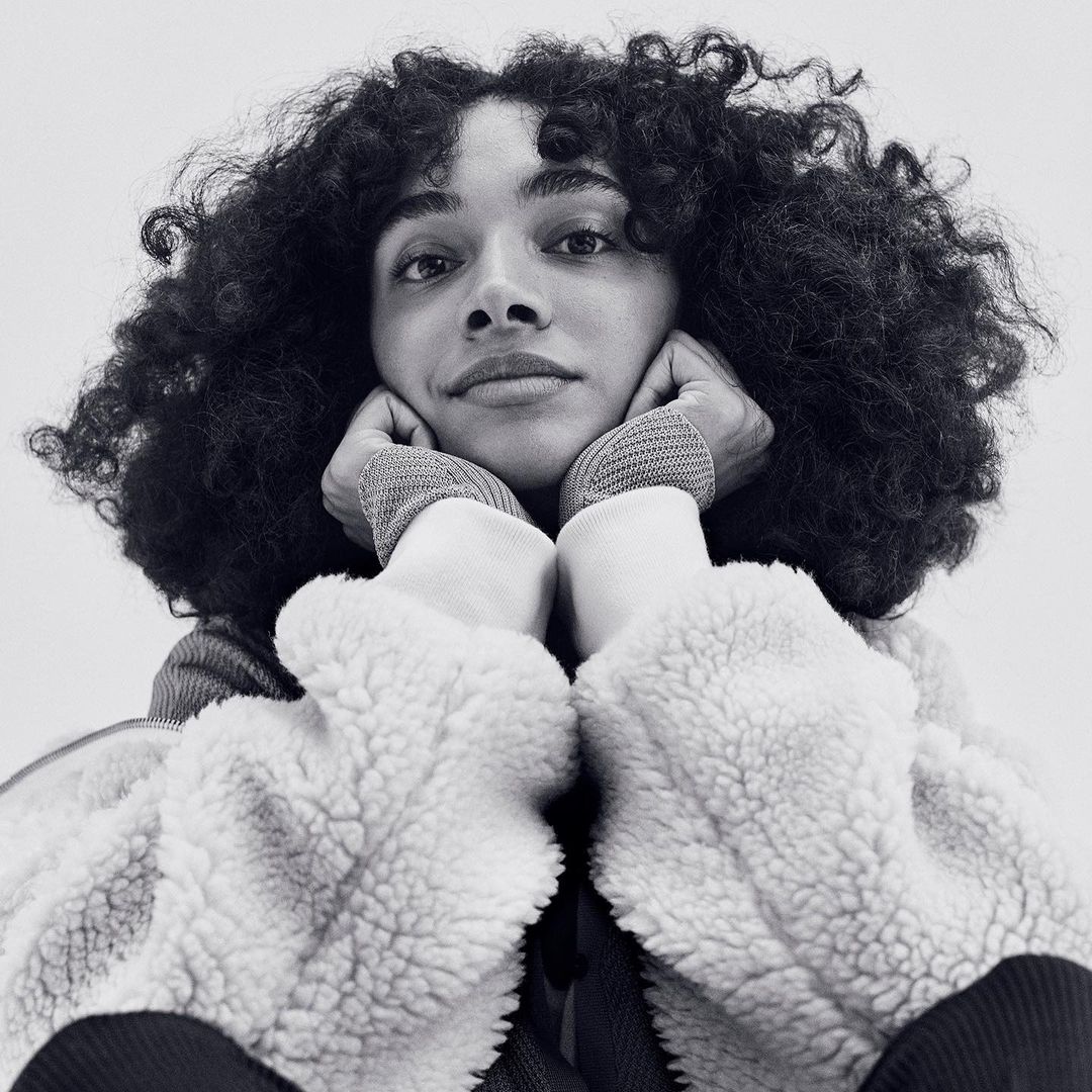 A young adult female with curly dark hair rests her face in her hands in a close-up, black and white studio portrait.