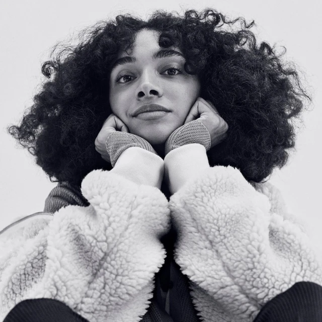 A young adult female with curly dark hair rests her face in her hands in a close-up, black and white studio portrait.
