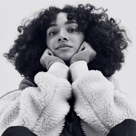 A young adult female with curly dark hair rests her face in her hands in a close-up, black and white studio portrait.