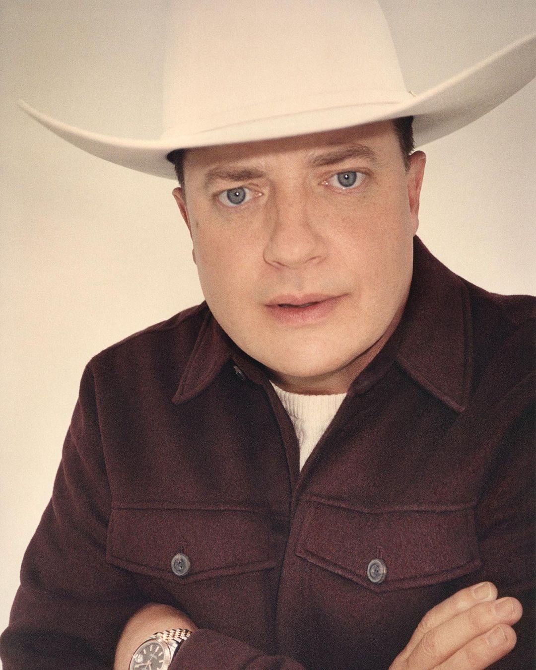 Portrait of actor Brendan Fraser wearing a white cowboy hat and a maroon shirt against a white backdrop in a studio setting.