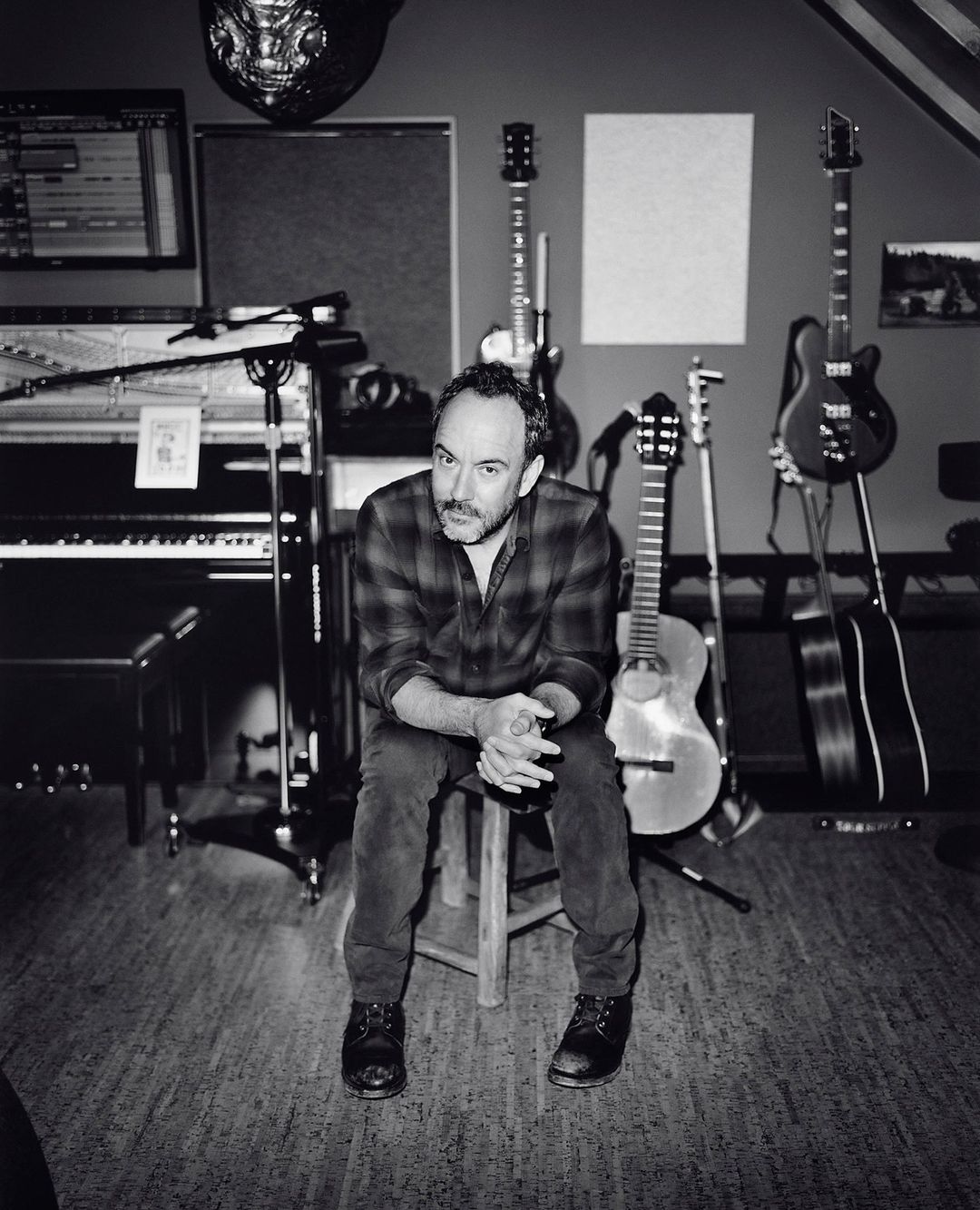Monochrome portrait of an adult man sitting on a stool in a music studio surrounded by instruments, including guitars and a piano.