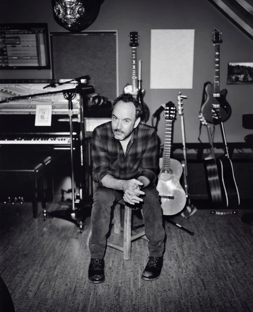 Monochrome portrait of an adult man sitting on a stool in a music studio surrounded by instruments, including guitars and a piano.