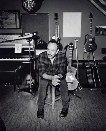 Monochrome portrait of an adult man sitting on a stool in a music studio surrounded by instruments, including guitars and a piano.