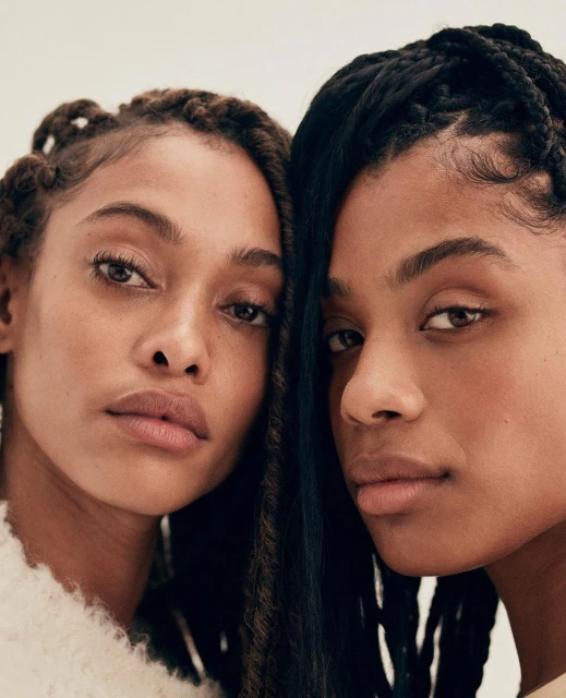 Close-up portrait of two beautiful Black women with distinctive braids, looking directly at the camera against a soft, neutral background.