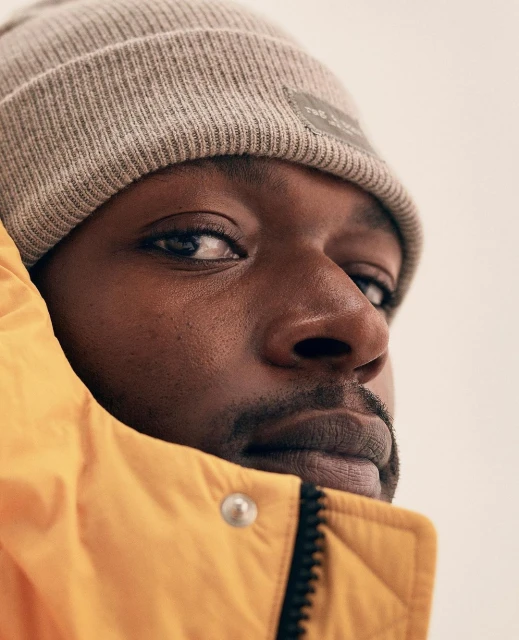 Close-up portrait of an African American adult male in a yellow puffer jacket and grey beanie hat, against a neutral backdrop.