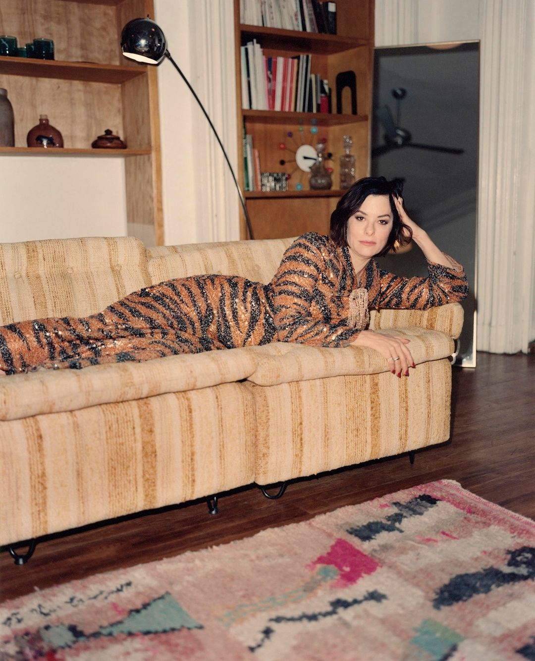 Adult woman in a tiger-print sequined dress lying on a striped couch in a warmly lit interior with books and a patterned rug.