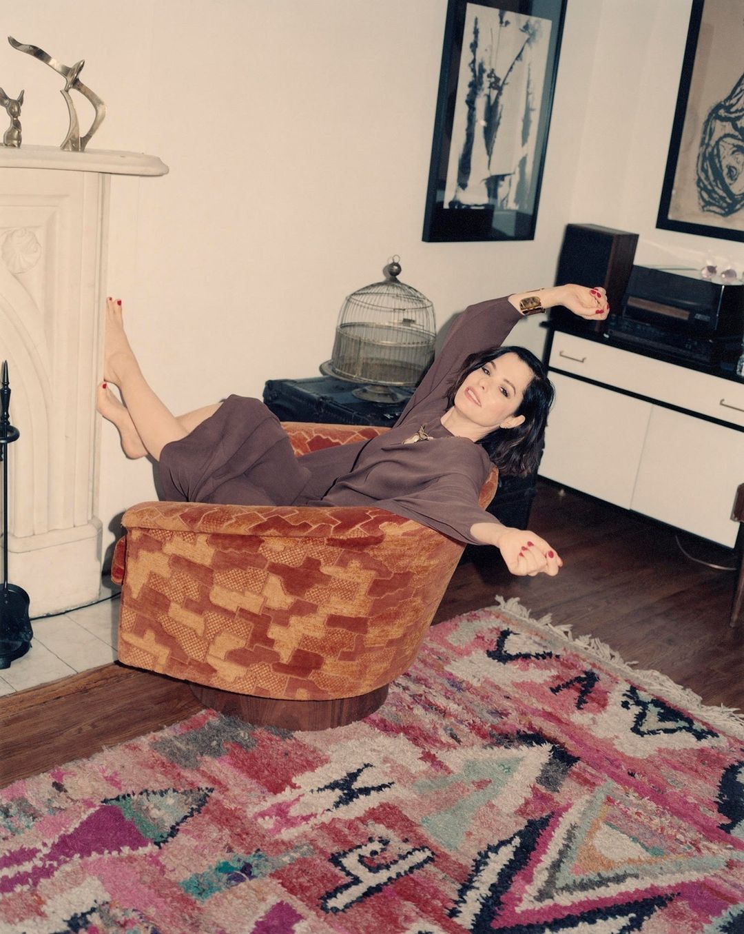 Woman lounging in an orange armchair in a living room setting. The room features a fireplace, artwork, and a patterned rug.