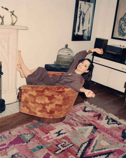 Woman lounging in an orange armchair in a living room setting. The room features a fireplace, artwork, and a patterned rug.