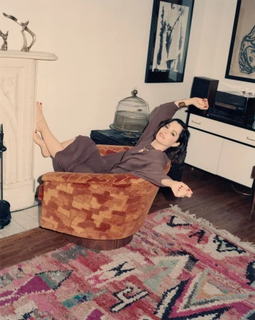 Woman lounging in an orange armchair in a living room setting. The room features a fireplace, artwork, and a patterned rug.
