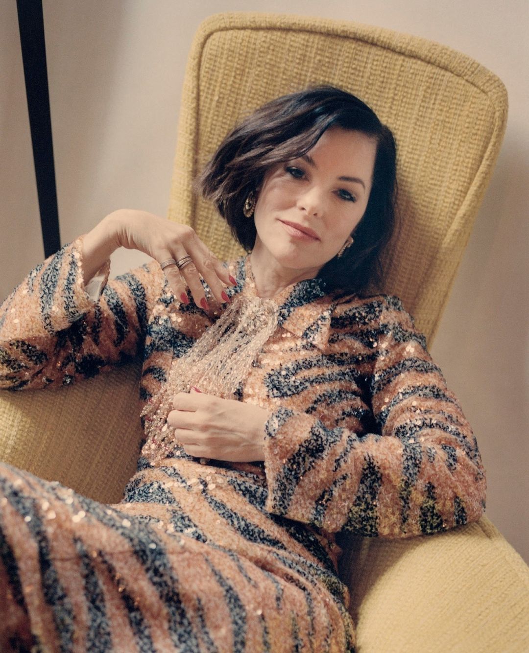 Portrait of woman with short dark hair in a yellow chair, wearing a sequined animal-print jacket and pants in an indoor setting.