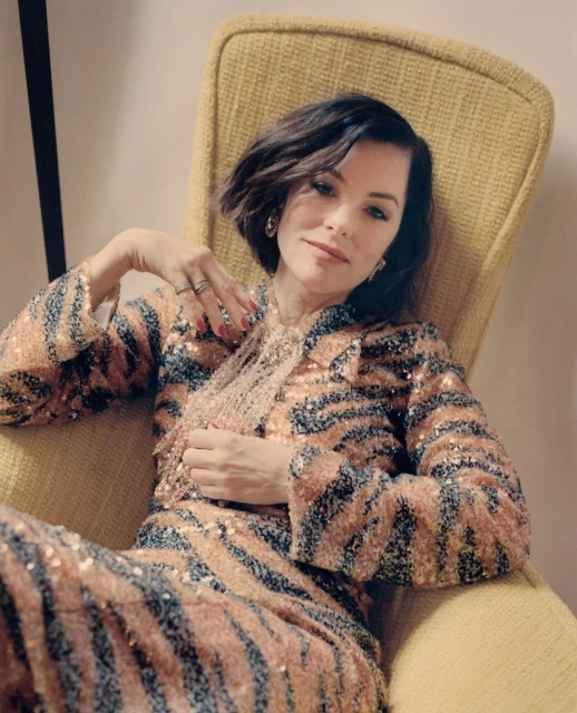 Portrait of woman with short dark hair in a yellow chair, wearing a sequined animal-print jacket and pants in an indoor setting.