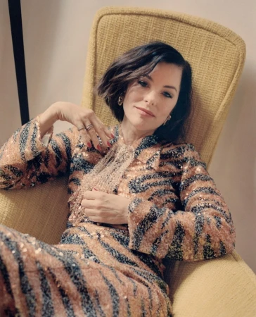 Portrait of woman with short dark hair in a yellow chair, wearing a sequined animal-print jacket and pants in an indoor setting.