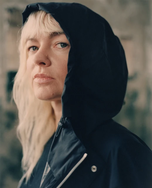 Portrait of a blonde adult female wearing a black hooded jacket, looking directly at the camera against a blurred urban backdrop.