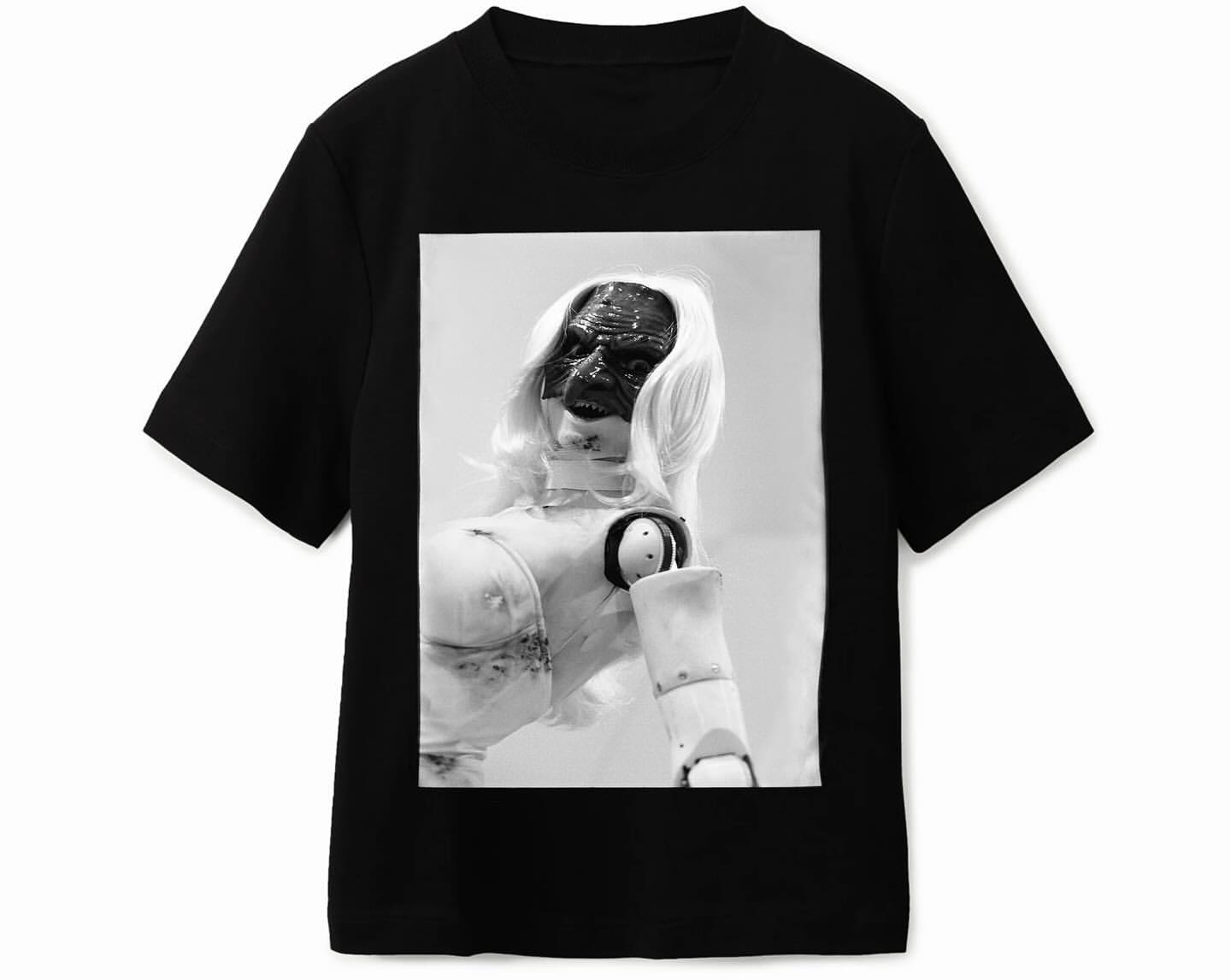 Black T-shirt with a black and white graphic of a mannequin-like figure with blonde hair and a dark face, centered on the front.