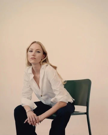 Blonde woman seated on a green chair wearing a white button-down shirt and dark trousers against a plain light background